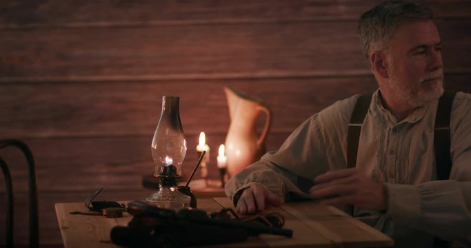 A mature pioneer type man sitting in a candle lit cabin turns to talk to his wife who hands him a Bible as she enters the scene then sits as he begins to read to her.