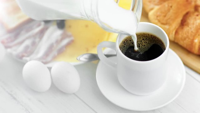 Pouring milk from jug into cup with black americano coffee during breakfast close-up