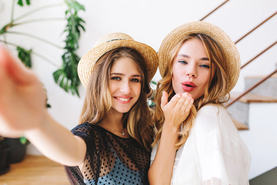 Long-haired girl with sincere smile making selfie while her friend posing with kissing face expression. Close-up portrait of two excited young ladies wearing hats and taking picture in room with plant