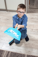 Uśmiechnięty chłopiec w okularach klęczy na podłodze i trzyma szufelkę wypełnioną papierkami   - obrazy, fototapety, plakaty