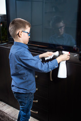 Chłopiec podczas sprzątania pokoju. Nanosi detergent na mebel za pomocą aplikatora. W drugiej...