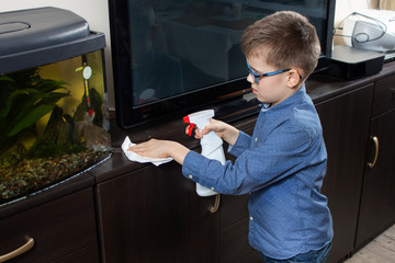 Chłopiec w niebieskiej koszuli i okularach sprząta w pokoju. Wyciera białą ściereczką kurze. © Tomasz