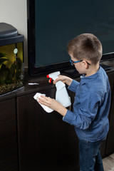 Chłopiec podczas sprzątania pokoju. Nanosi detergent na mebel za pomocą aplikatora. W drugiej ręce trzyma białą ściereczkę do wycierania. - obrazy, fototapety, plakaty