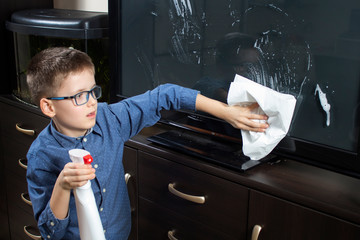 Chłopiec w niebieskiej koszuli i okularach sprząta w pokoju. Wyciera białą ściereczką kurze.
