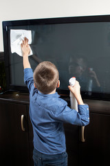 Chłopiec w okularach podczas sprzątania. Wyciera ściereczką ekran telewizora z kurzu. - obrazy, fototapety, plakaty