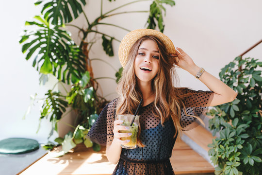 Attractive long-haired girl in hat with nude makeup standing with cocktail in room with big plants in pots. Indoor portrait of glad young woman in wristwatch holding glass of juice.