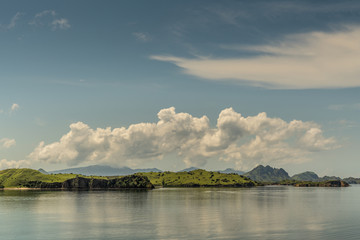 Rinca Island, Indonesia - February 24, 2019: Flat islets off Westside coast in Savu Sea under cloudscape with white patches. Green hills in distance and flat sea.