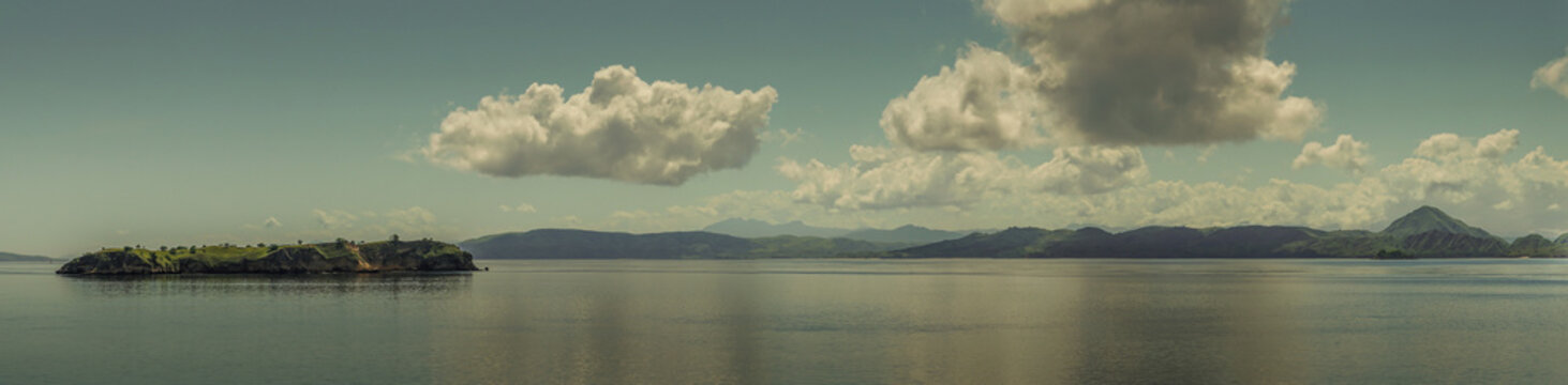 Rinca Island, Indonesia - February 24, 2019: Panorama shot of Westside coast in Savu Sea under cloudscape with white and darker patches. Green hills and flat sea. Islet up front.
