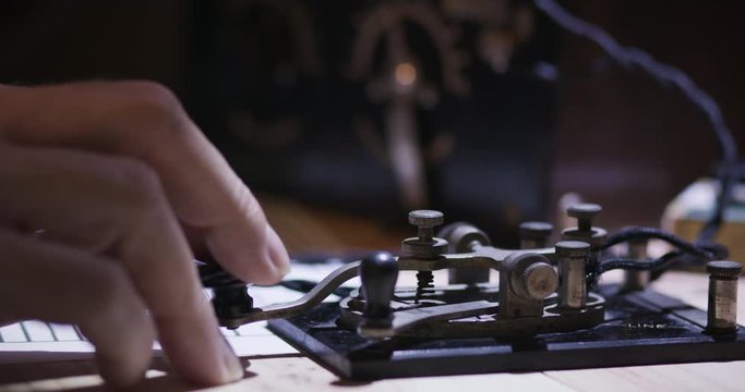 An operator picks up a pair of headphone from a workstation then starts transmitting in Morse Code on a vintage code key circa WWII