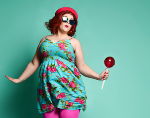 Funny overweight fat chubby woman in funny hat and colorful clothes and extra big lollipop tries to...