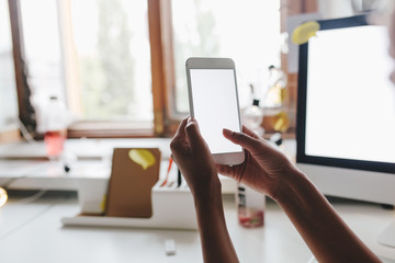 Photo of smartphone with white screen in female hands with big window on background.  Woman with dark skin using her phone in room with computer.