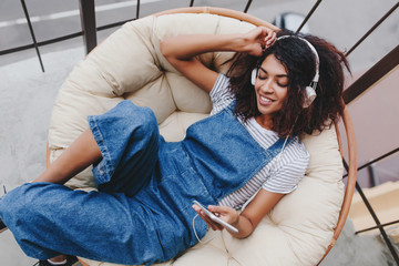 Blissful black young woman in vintage denim clothes chilling on terrace in white headphones....