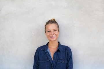 Young millennial girl is standing over the grey concrete wall in jeans dungree.
