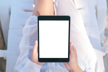 Top view mockup image of a woman holding and using a black tablet pc with blank desktop screen while laying down on beach chair on the beach