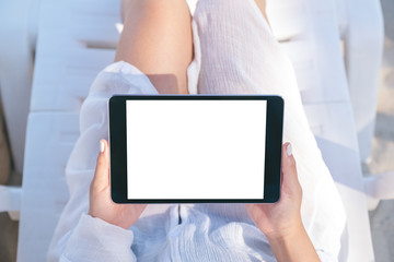 Top view mockup image of a woman holding and using a black tablet pc with blank desktop screen while laying down on beach chair on the beach