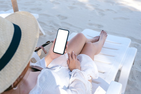 Mockup image of a woman holding white mobile phone with blank desktop screen while laying down on beach chair on the beach