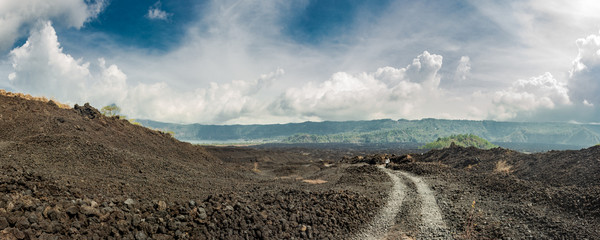 Walking tourist on a dirt road leading to green hills among black lava and dramatic cloudy sky. Panoramic view of the slopes of the Batur volcano covered with black lava. © everigenia