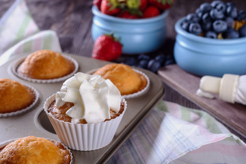 Homemade freshly baked muffins and one of them is decorated whipped cream on top are in baking dish on a wooden table next to cream and berries for decorating
