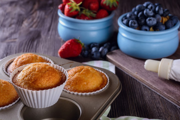 Homemade freshly baked muffins in baking dish on a wooden table next to cream and berries for decorating