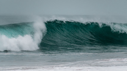 A large barreling wave crashing onto the shore spraying mist