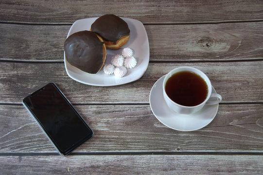 A cup of black tea on a saucer and a plate with two donuts in chocolate icing and pieces of bize, are lying on a wooden table wall next to a smartphone. Coffee break. Close-up.