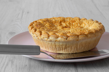 Steak and ale pie cooked on a pink plate with a stainless steel serving utensil on a grey wood background