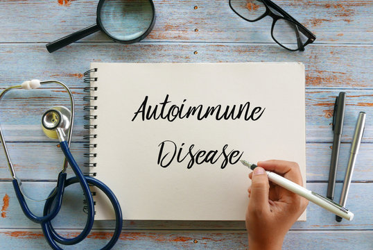 Top view of magnifying glass,glasses,stethoscope,pen and hand writing ' Autoimmune Disease ' on notebook on wooden background,
