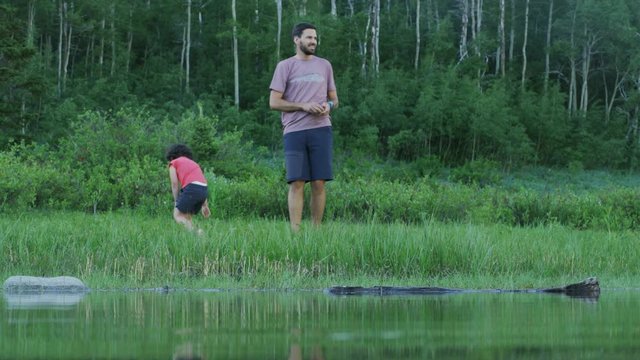 Camera Looks Through Lake At Father Teaching Young Daughter To Skip Rocks, Son Enters Frame And Throws Rocks