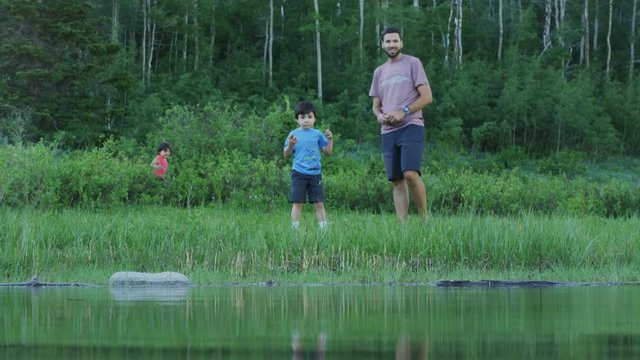 Camera Looks Over Lake At Father And Son Skipping Rocks, Rock Splashes Near Camera Lense