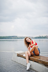 Sexy curly model girl in red top, jeans denim shorts, eyeglasses and sneakers posed on bench against lake.