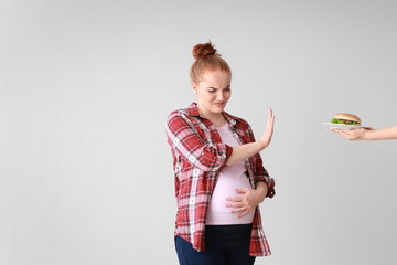 Young pregnant woman refusing to eat tasty burger on light background