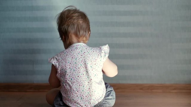 Little cute baby girl sits on the floor and turns away from the camera in the sun rays on the background of beautiful blue wallpaper in slow motion. Sunbeam is visible on the wall