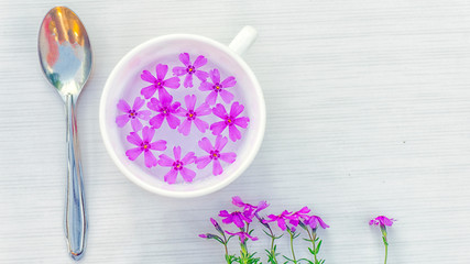 White cup of green tea with purple flowers on a white table. Nearby is a teaspoon and fresh flowers. Top view.