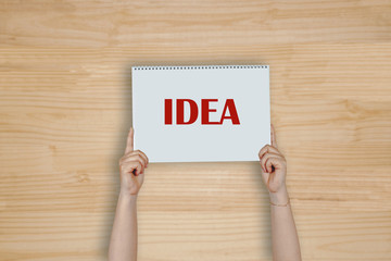 Woman hands holding high a post card that says IDEA against a wood board.