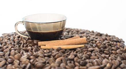A cup of coffee on a white table, around a cup scattered grains of roasted coffee and fresh cinnamon