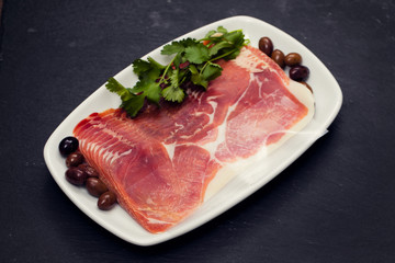 prosciutto with olives and parsley on white dish on black ceramic background