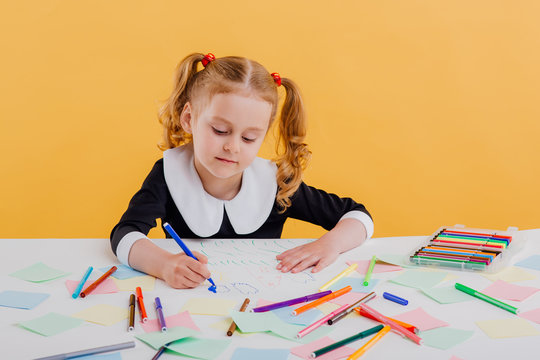 beautiful little girl draws with blue flamaster on the white table, dressed in school uniform sitting at the white table, isolated on yellow background, copy space,