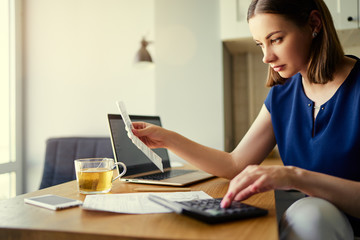 Thoughtful young woman using a laptop computer sitting at her kitchen holding utility bill and bank statements. Home interior.