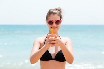 Young woman is holding a spray of sun cream for body at the camera at the beach