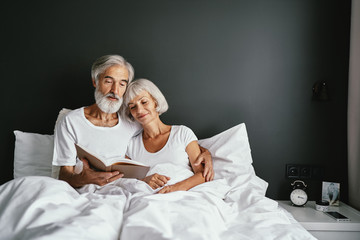 Happy family. Cozy home. Beautiful senior woman and her husband reading book together in bedroom. - Powered by Adobe