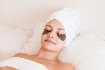 Fototapeta na wymiar Beautiful young woman with under eye patches in bathrobe lying in bed. Happy girl taking care of herself. Beauty skincare and wellness morning concept