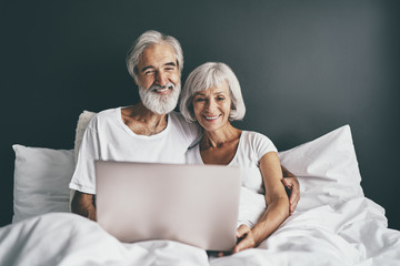 Happy family. Cozy home. Beautiful senior woman and her husband using laptop computer together in bedroom.