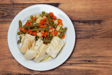 boiled cod fish with boiled vegetables on white plate on wooden background