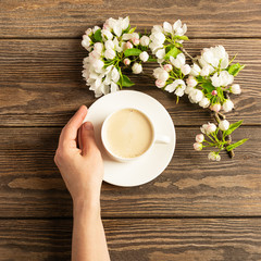 A cup of hot aromatic coffee in female hands and a blooming apple tree branch on a wooden background. Flat layout.