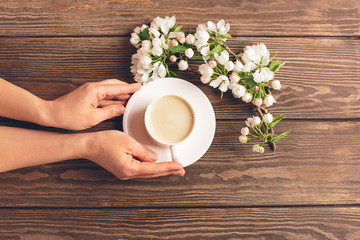 A cup of hot aromatic coffee in female hands and a blooming apple tree branch on a wooden background. Flat layout.