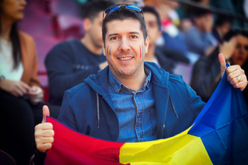 Happy supporter holding Romanian national flag in hands at international sport event – man in the stands of stadium cheering favourite team – soccer fan in arena with blurred people in background