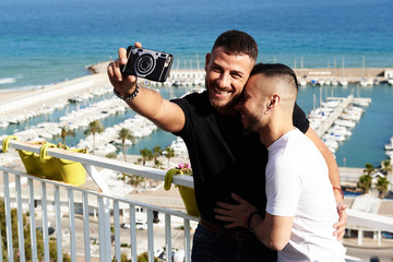 Una pareja gay se hace un selfie en el balcón con el puerto detrás de ellos en un día de verano