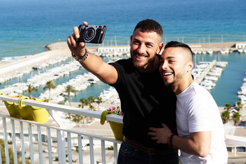 Una pareja gay se hace un selfie en el balcón con el puerto detrás de ellos en un día de verano