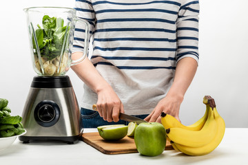 cropped view of woman cutting apple near blender and bananas on white