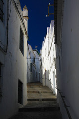 Low angle view on narrow empty alley with facades of white houses and steps upstairs contrasting with dark blue sky in traditional spanish village Arcos de la Frontera - Andalusia, Spain